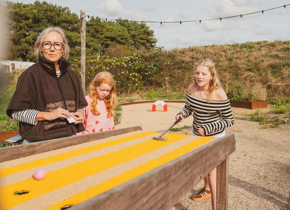 Grandmother and two girls playing mini golf on a yellow table outdoors at a holiday park on a sunny day.