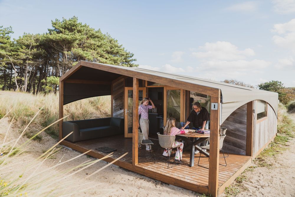Family enjoying outdoor dining at a modern wooden beach cabin surrounded by sand dunes and pine trees.