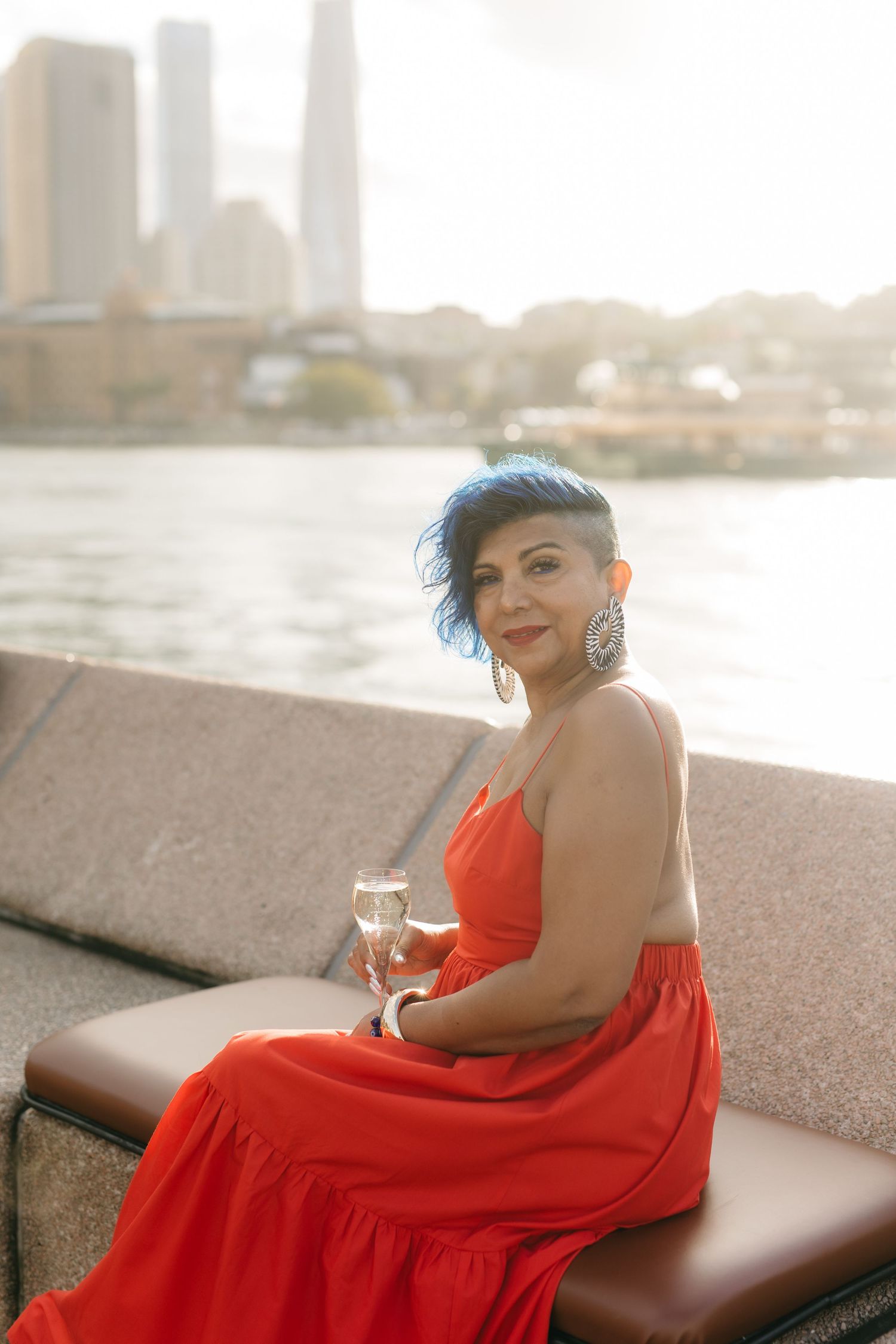 Woman in red dress holding champagne glass seated by waterfront with city skyline backdrop.