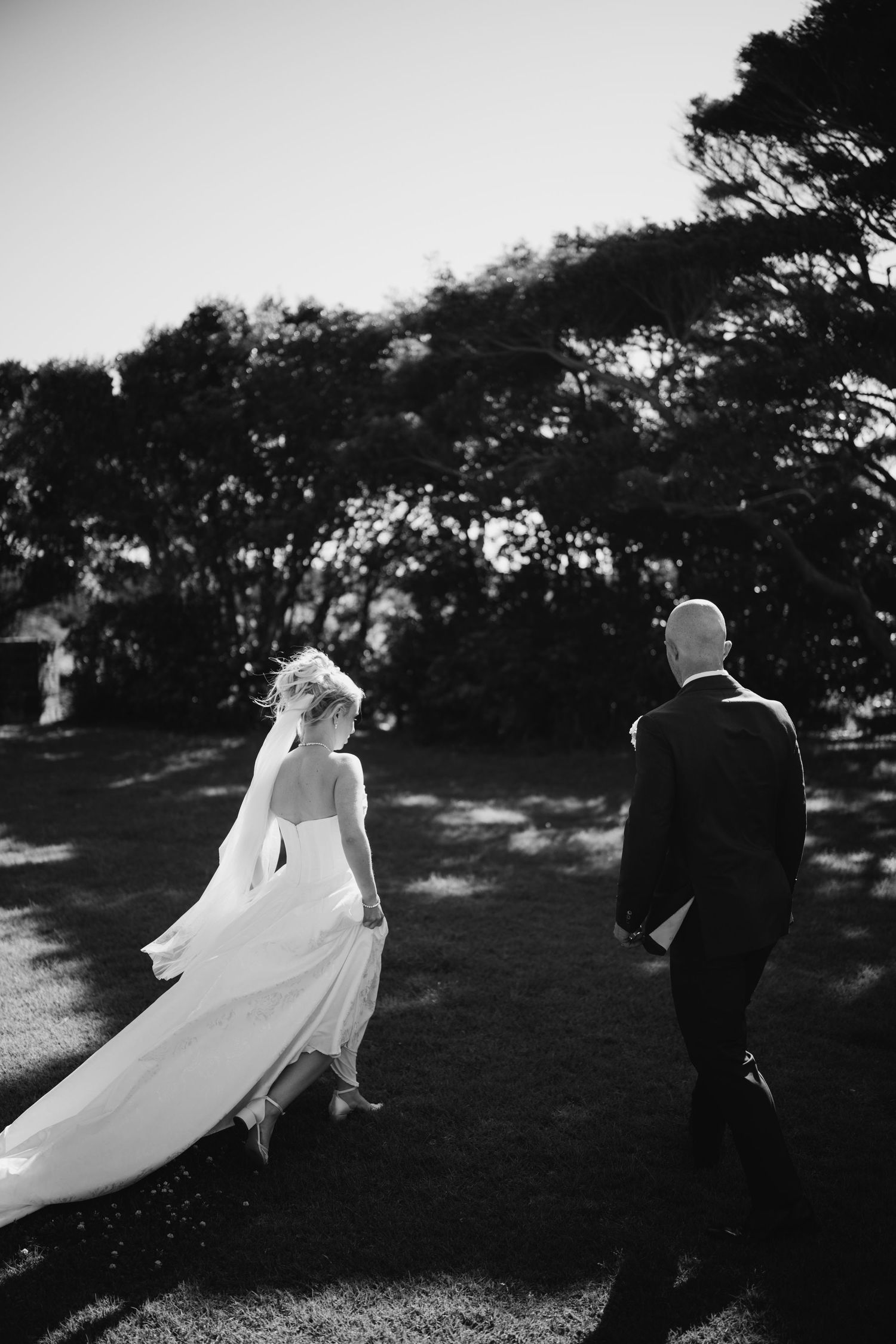 Bride and groom share a romantic moment on a sandy beach, she holds a burgundy bouquet in a white gown, he wears a black tuxedo.