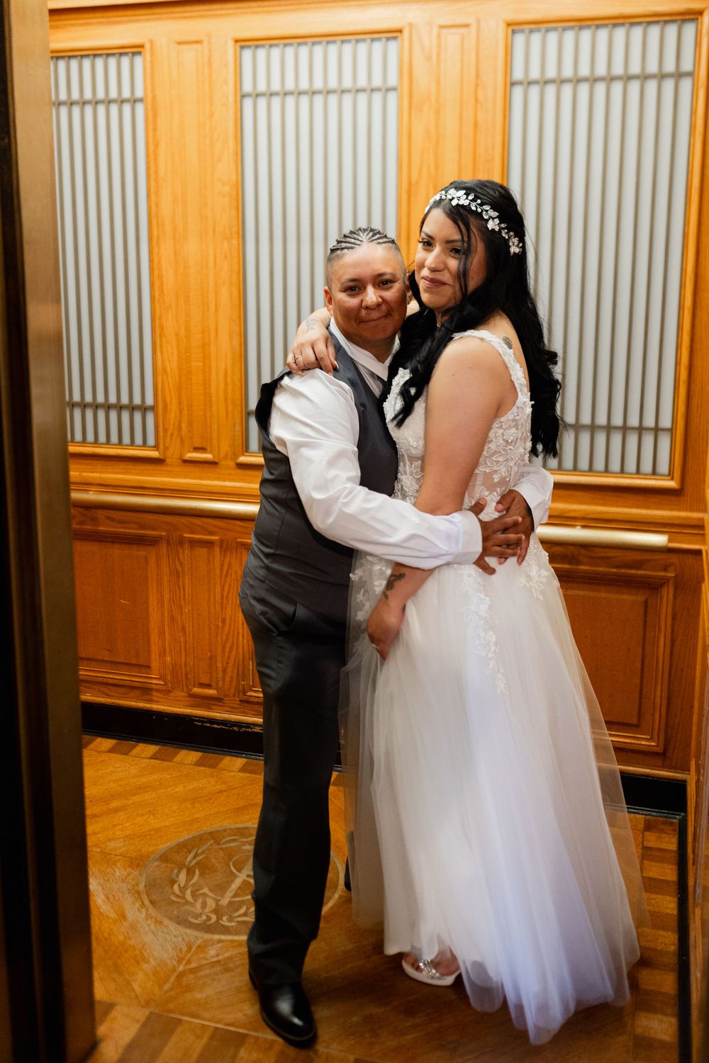 Two brides embrace in an elevator, one in a white wedding gown with floral headpiece, the other in a gray vest suit.