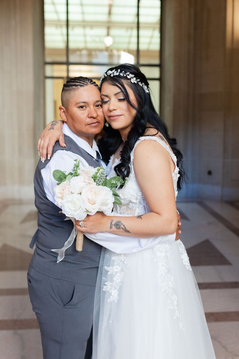 Two brides embrace tenderly in elegant wedding attire, one in a white lace gown with floral crown, the other in a gray suit.