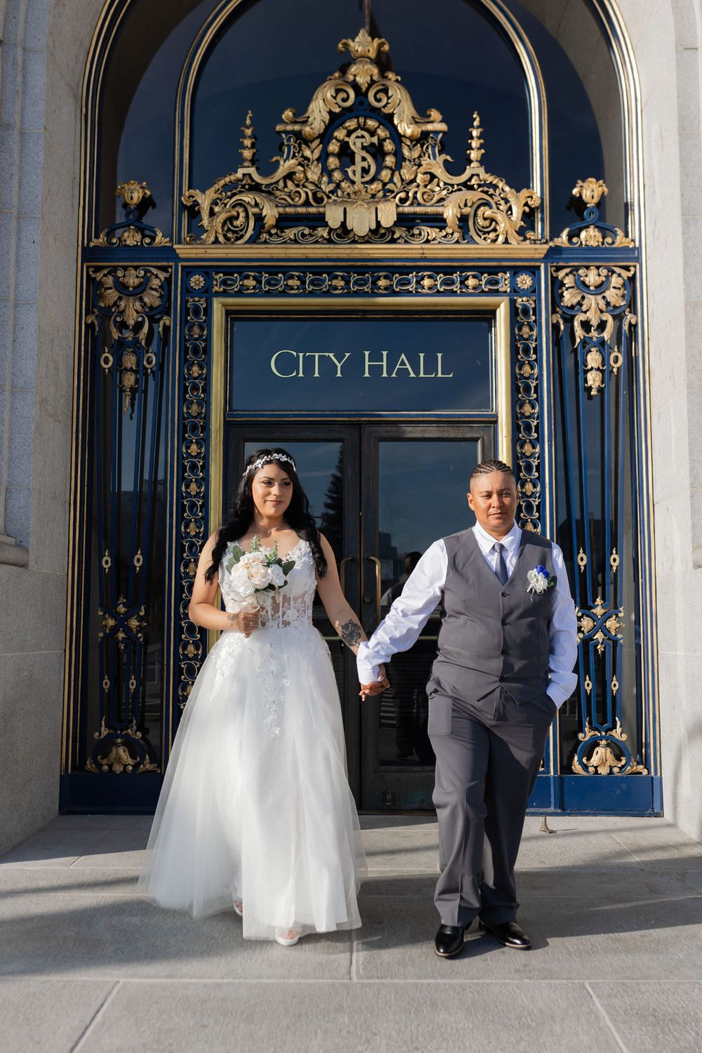 Newlywed couple holding hands outside ornate City Hall entrance, bride in white gown with bouquet, groom in gray vest.