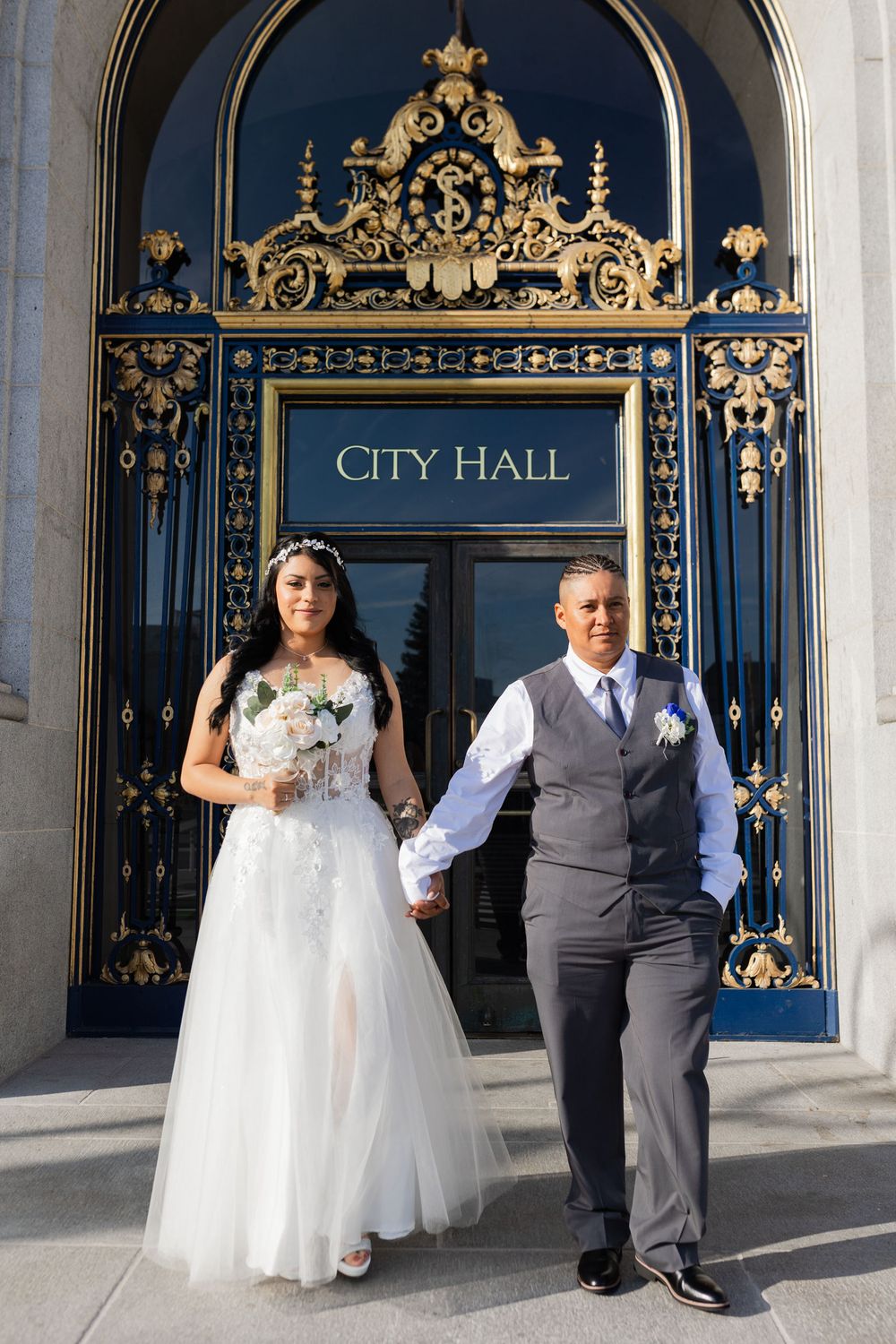 Newlywed couple holding hands in front of ornate City Hall golden gates, bride in white gown holding bouquet, groom in gray suit.