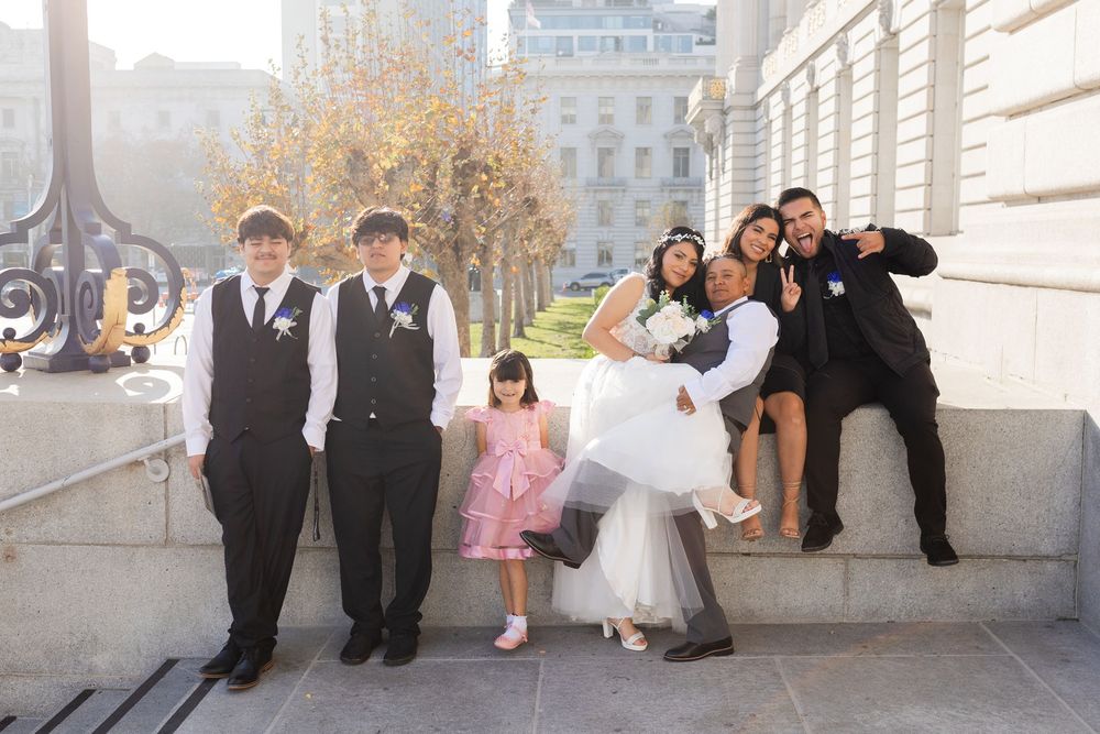 Wedding party poses on steps outside a grand building, featuring bride, groom, groomsmen, and flower girl on a sunny day.