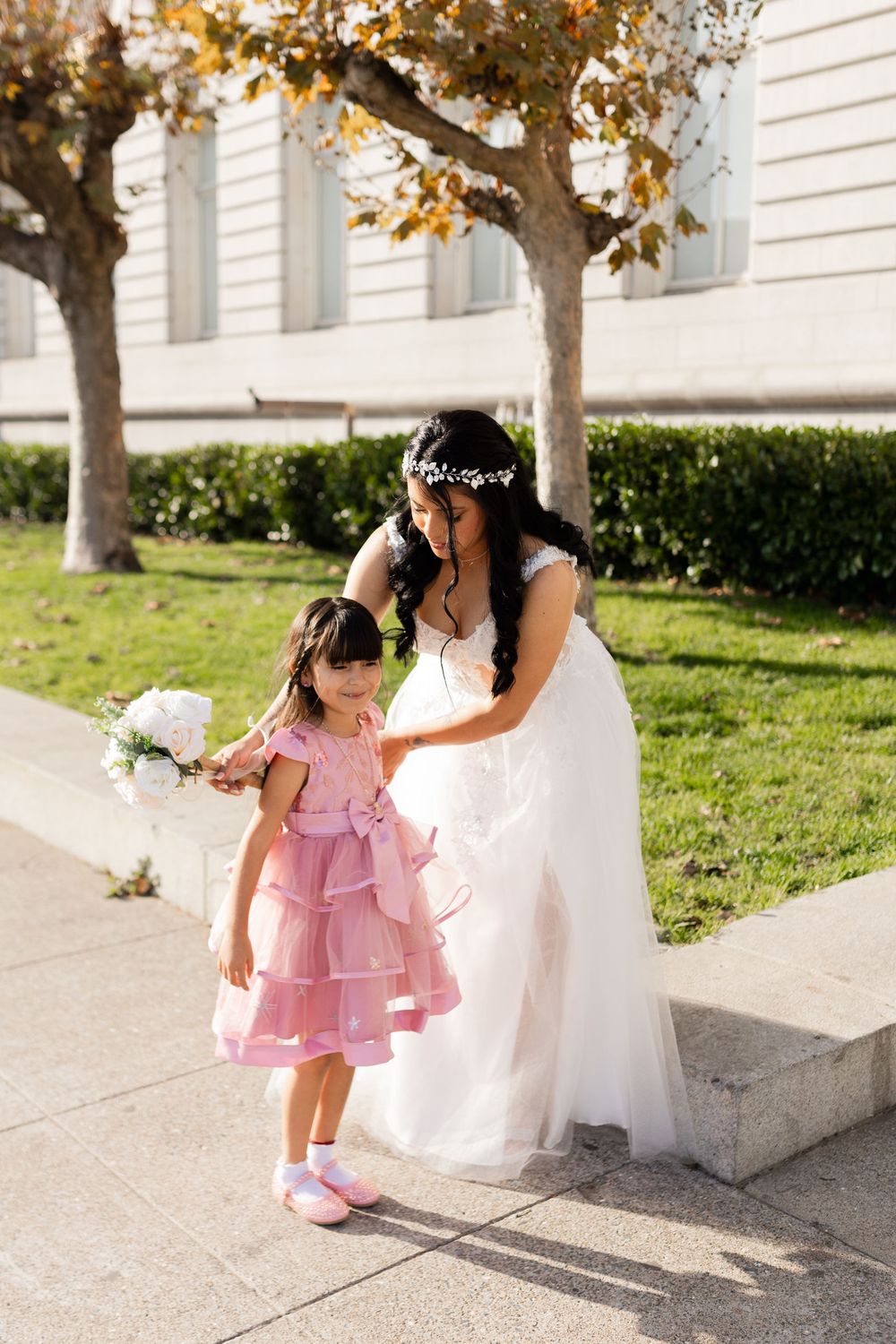 Bride in white gown kneels beside flower girl in pink dress outdoors near City Hall.