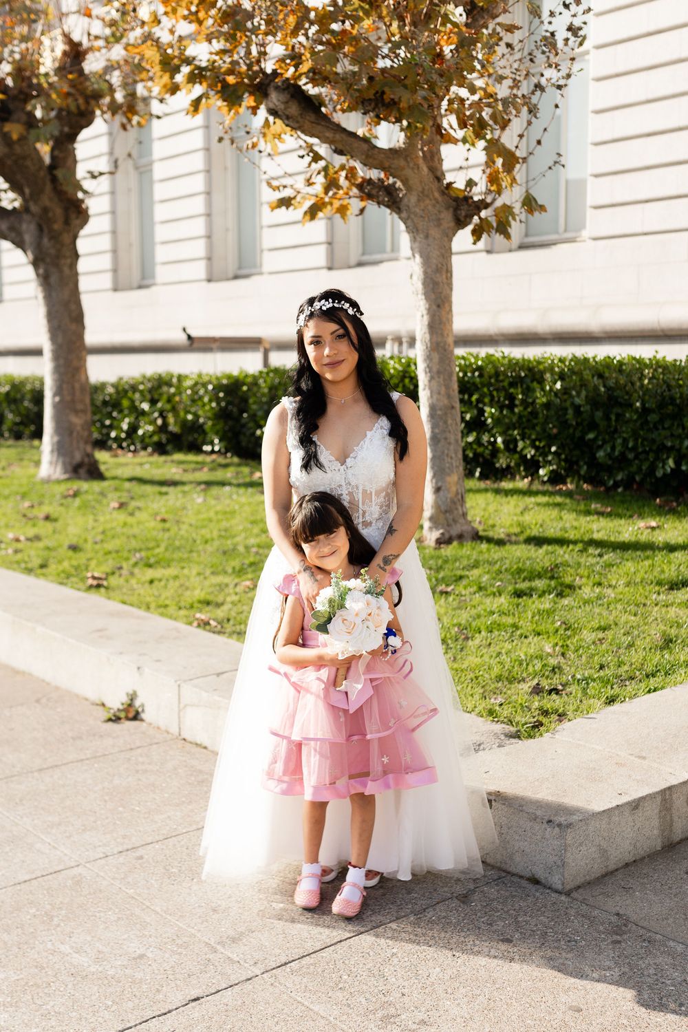 Bride in white gown with flower girl in pink dress holding bouquet, posing outdoors near trees and green hedges.
