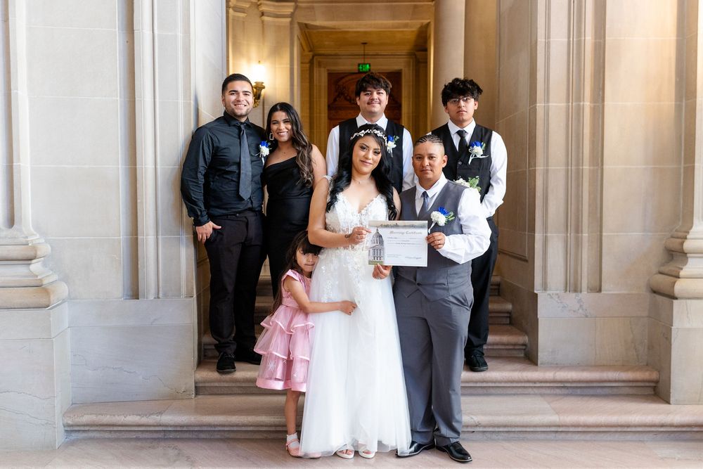A joyful wedding party poses on elegant stone steps, with the bride in white holding a marriage certificate alongside her groom and family.