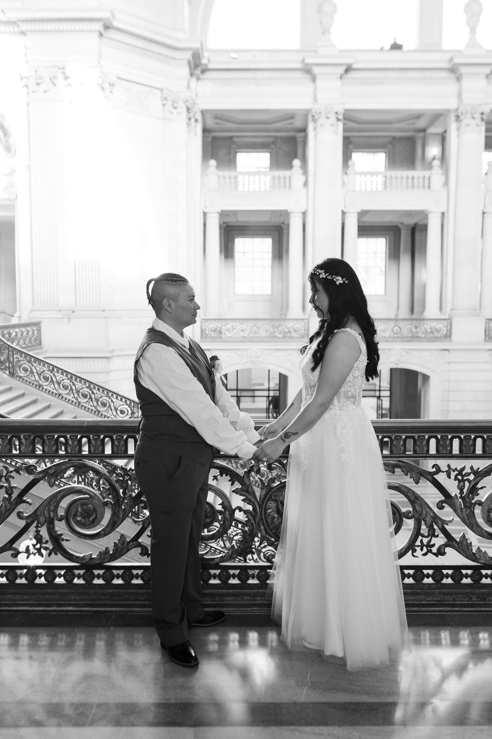 Black and white photo of a couple exchanging vows at an ornate indoor venue, holding hands in wedding attire.
