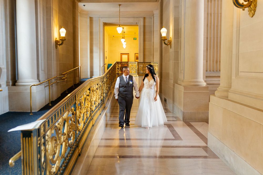 Bride and groom walking hand in hand through an elegant gold-railed hallway at San Francisco City Hall.