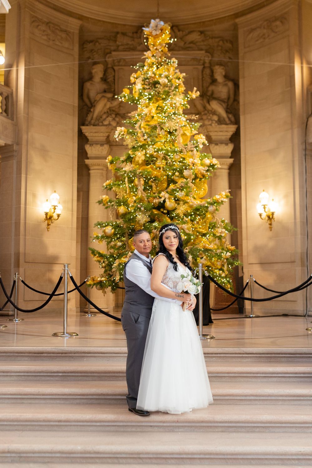 Couple poses in wedding attire before a glowing gold Christmas tree in an elegant grand hall.