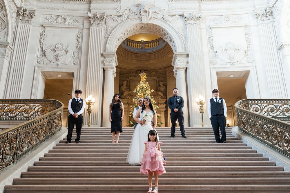 Wedding party poses on grand staircase of ornate venue with flower girl in pink dress in foreground.