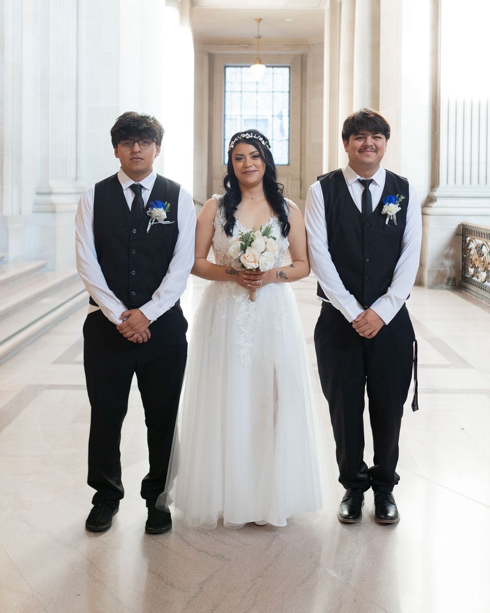 Young woman in white wedding gown holding bouquet flanked by two men in black vests and ties in elegant hall.