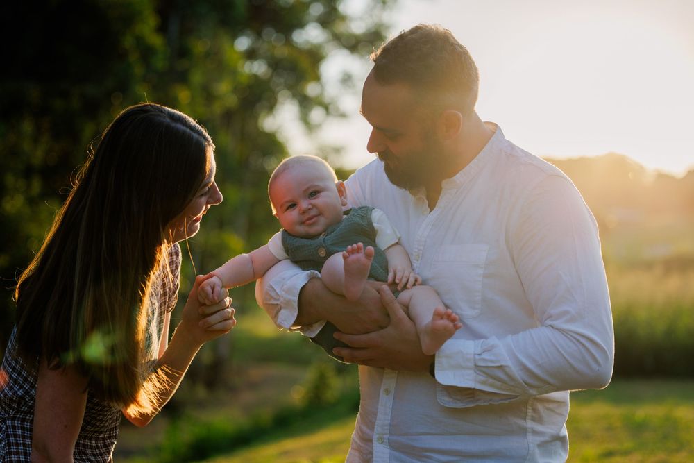 A smiling baby is held by a bearded father in a white shirt while the mother looks on in warm golden sunset light.