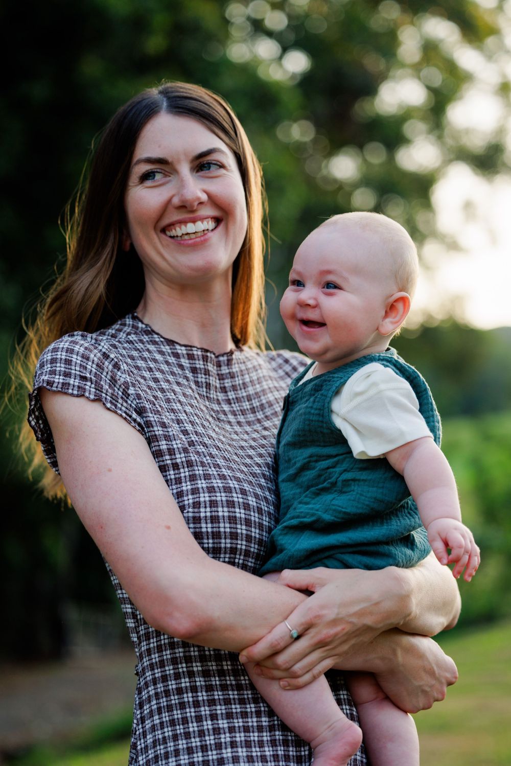 A laughing woman in a checkered dress holds a smiling baby in a green outfit outdoors at golden hour.