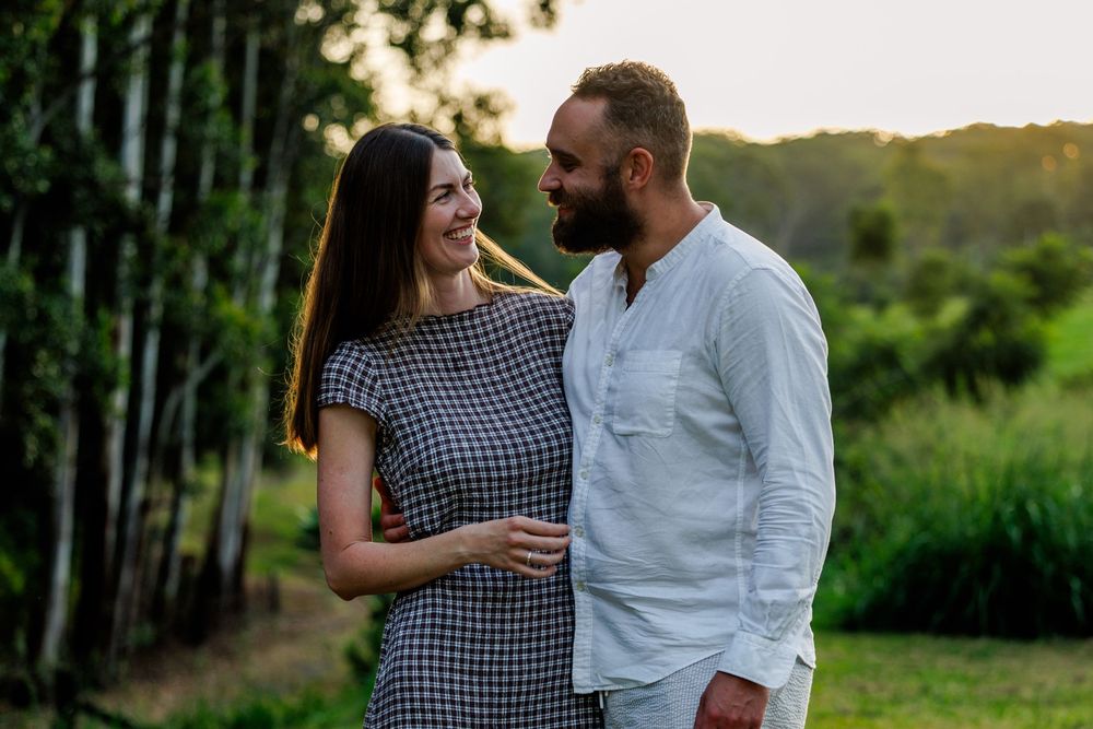 A smiling couple embraces outdoors in warm evening light with trees and green fields behind them.