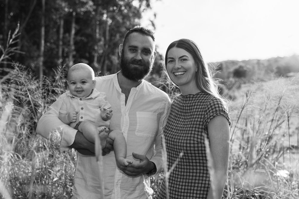 A black and white family portrait of a bearded father holding a baby beside the smiling mother in a field of tall grass.