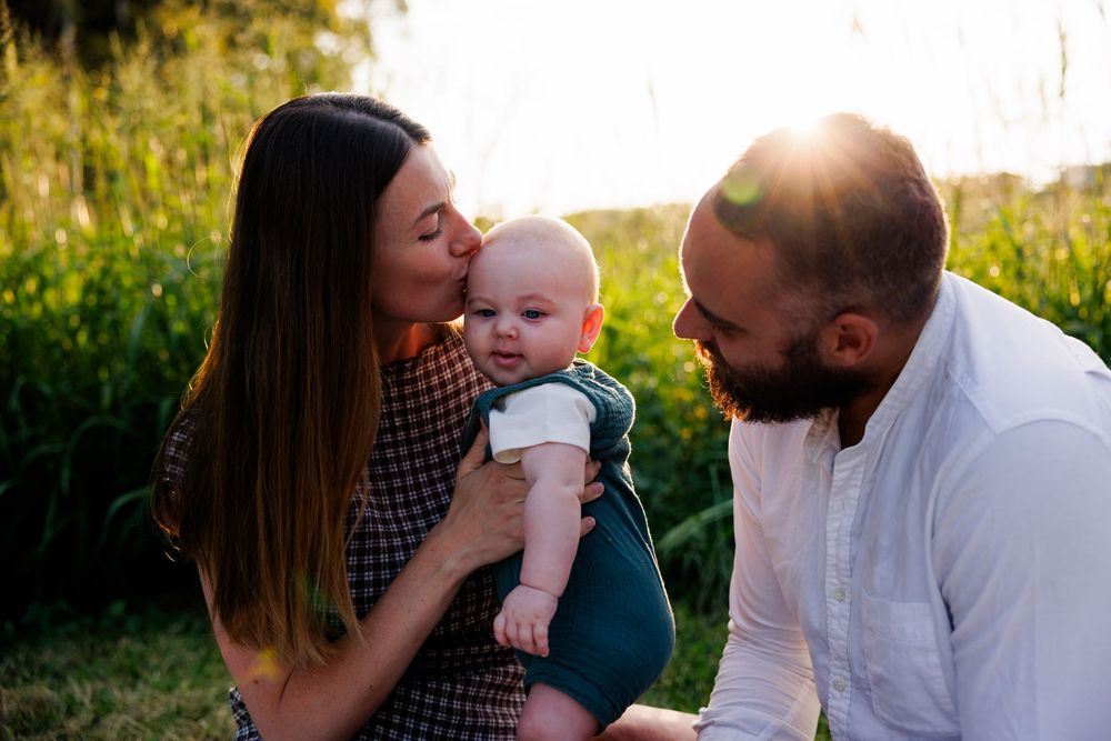 A mother kisses her baby's cheek while the father sits beside them in a sunlit grassy field with a warm sun flare.
