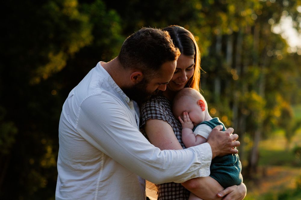 A father and mother hug their baby close in a tender family embrace beneath trees in soft golden light.