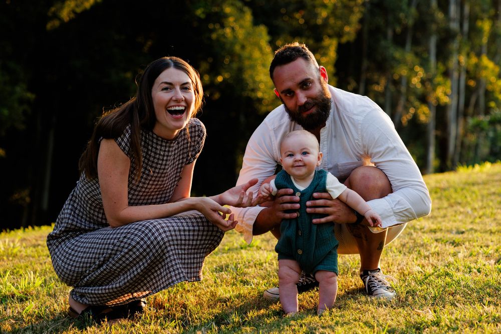 Smiling couple crouching on sunlit grass while holding their happy baby between them during a golden hour family photo session.