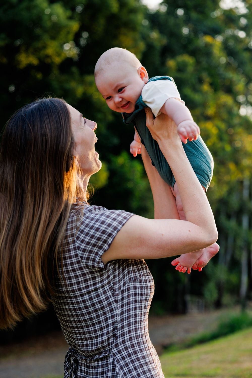 Mother lifting her smiling baby high in the air outdoors, with softly blurred green trees in the background.