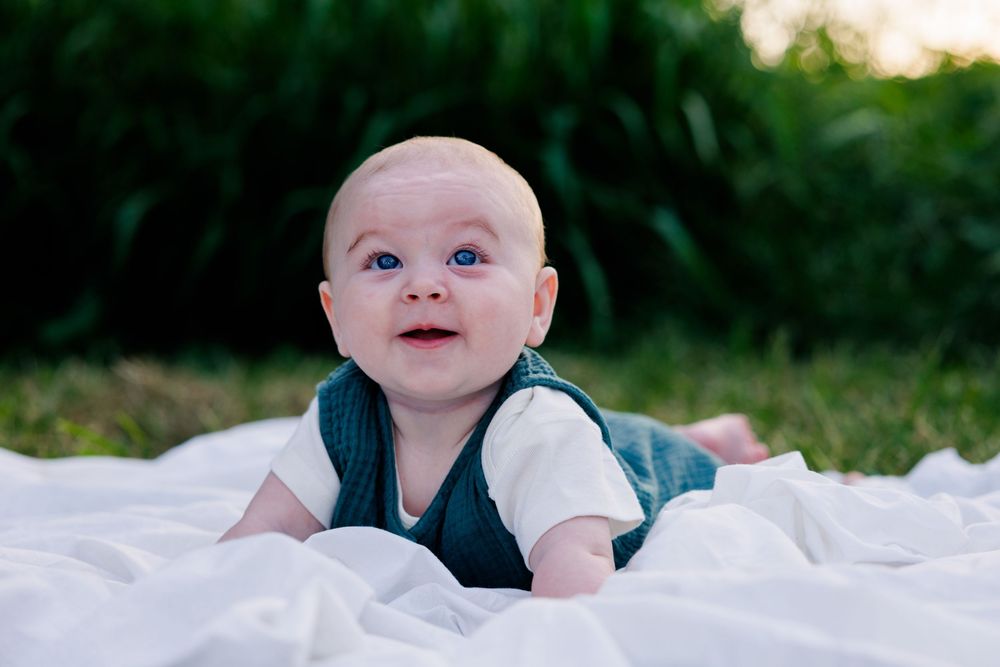Blue-eyed baby in a green vest lying on a white blanket on the grass, smiling at the camera in a natural outdoor setting.