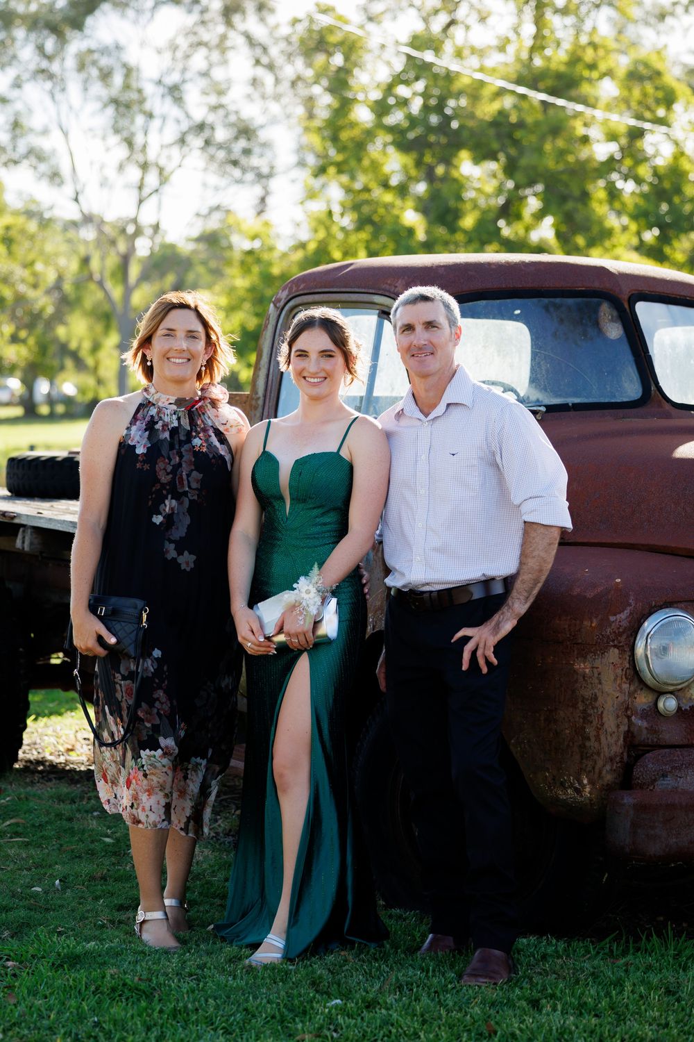 Woman, young woman in a green slit gown, and man pose in front of a rustic vintage truck on a grassy lawn.