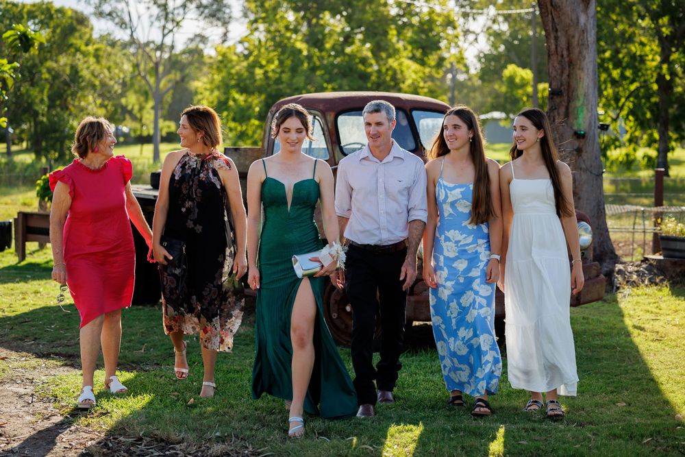 Group of women in formal dresses and a man walk across the grass toward a vintage truck on a sunny day.