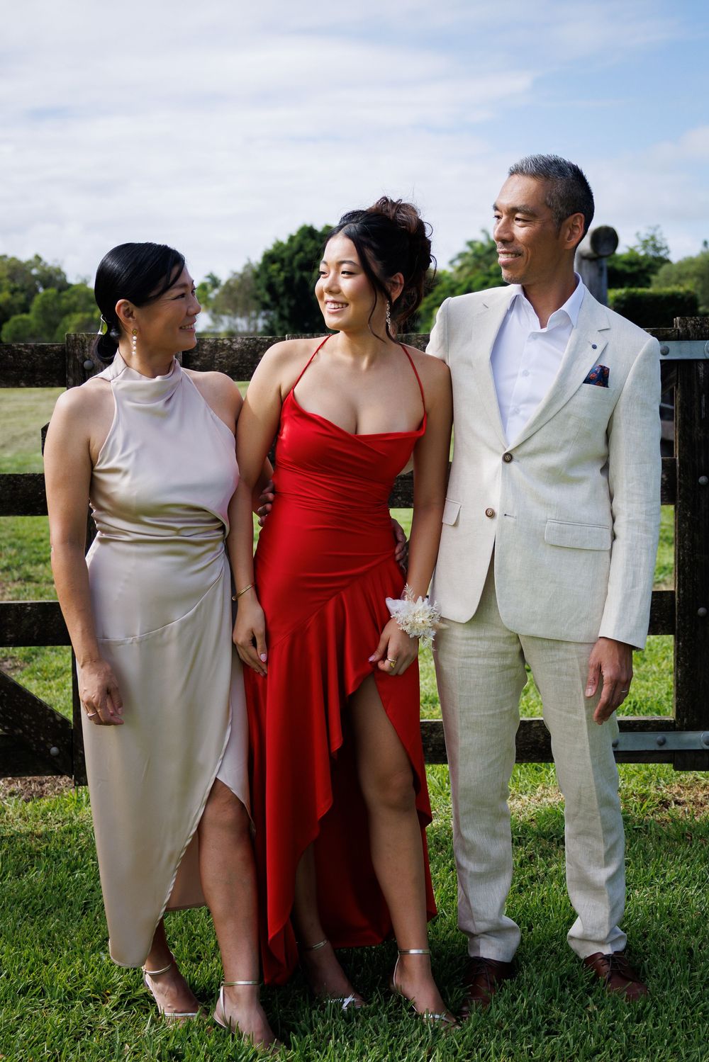Young woman in a red formal dress poses between two family members on grass in front of a wooden fence.