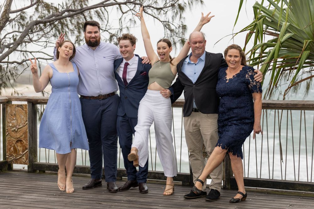 Six smiling wedding guests in semi-formal outfits pose on a wooden boardwalk with a coastal view and palm fronds behind them.