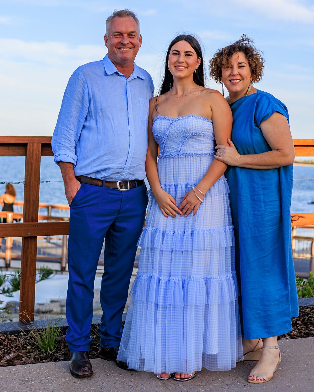 A smiling man and two women in blue outfits pose together on a seaside deck at sunset.