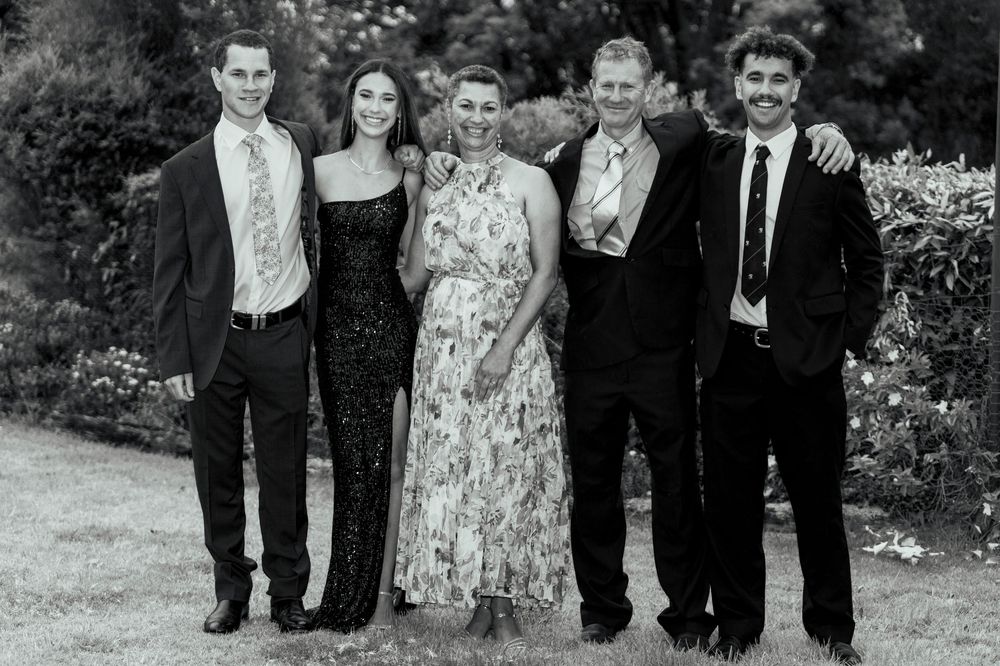 Five people in formal attire pose together on a grassy lawn surrounded by greenery in a black and white group portrait.
