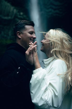 A smiling couple holds hands tenderly in front of a misty waterfall backdrop.