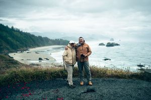 Couple posing on rocky Oregon coast overlook with scenic beach and sea stacks in cloudy background.
