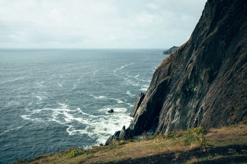 Rugged dark rocky sea cliffs plunging into choppy ocean waters under a dramatic overcast sky.
