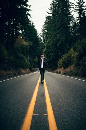 Man walking along center line of empty forest road flanked by tall evergreen trees.