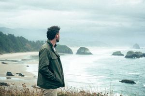 A man in a green jacket gazes at a dramatic rocky coastline under an overcast sky.