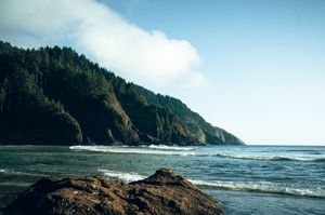 Lush forested coastal cliffs meet the Pacific Ocean waves under a partly cloudy blue sky.