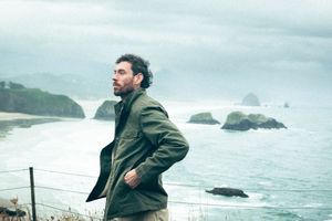 Bearded man in olive jacket standing on coastal cliff overlooking rocky Oregon coastline.
