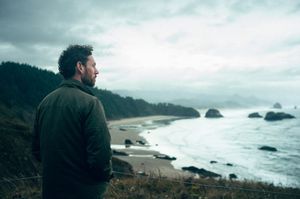 Man in green jacket gazing at dramatic overcast Oregon beach with sea stacks.