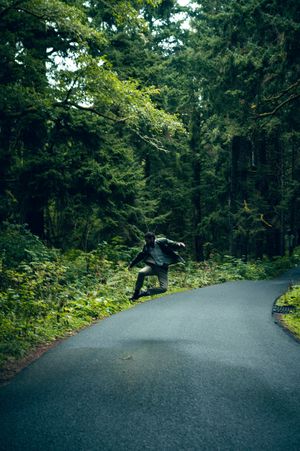 Person jumping mid-air on a winding road surrounded by lush green forest trees.
