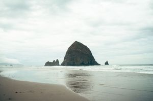 Iconic Haystack Rock rises from the sandy shore at Cannon Beach, Oregon under a cloudy sky.