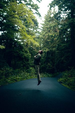 A man joyfully jumps on a winding road surrounded by towering green trees in a lush Pacific Northwest forest.