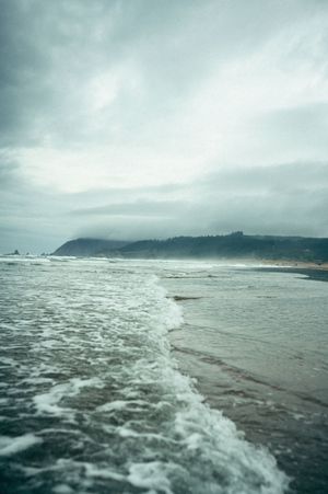 Dramatic overcast ocean waves washing onto a wide sandy beach with a misty headland in the background.