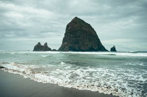 Waves crash near the base of Haystack Rock and sea stacks at Cannon Beach, Oregon on an overcast day.