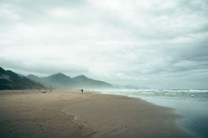 Two distant figures walk along a wide misty sandy beach with foggy mountain backdrop.