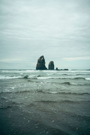 Two dramatic sea stack rock formations rising from choppy ocean waves under cloudy gray sky.