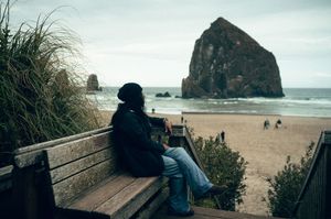 Person sitting on a wooden bench overlooking Haystack Rock and Cannon Beach, Oregon on a cloudy day.