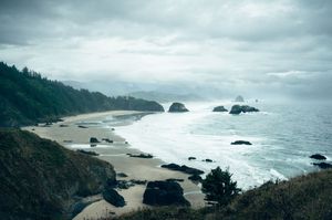 Dramatic overcast view of Oregon coastline with sea stacks, rocky beach, and misty forested headlands.