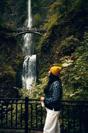 A woman in a yellow beanie and striped jacket admires Multnomah Falls waterfall and bridge in Oregon.